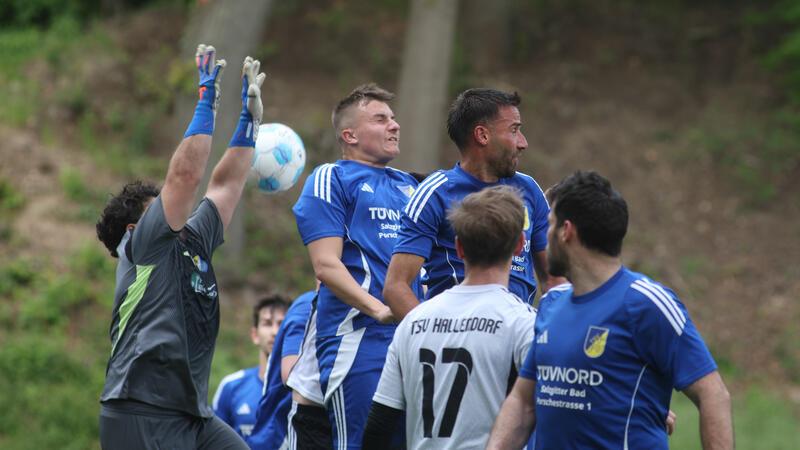 Fußballspieler in blauen Trikots und ein Torwart mit erhobenen Armen springen vor einem Spieler in weißem Trikot mit der Aufschrift TSV Hallendorf.