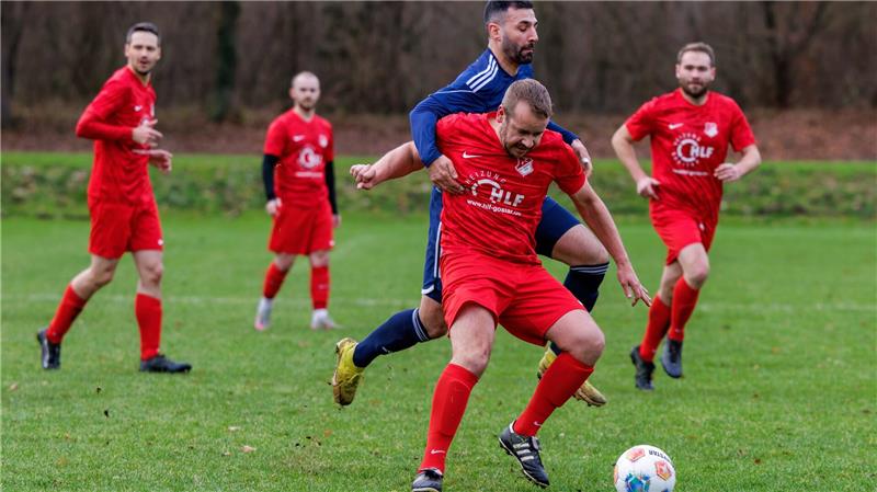 Fußballspiel mit mehreren Spielern, zwei im Vordergrund kämpfen um den Ball, einer trägt blau, der andere rot mit weißem Logo.