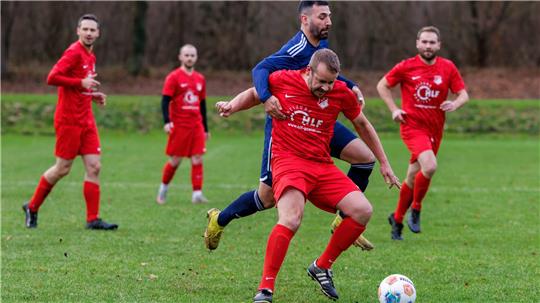Fußballspiel mit mehreren Spielern, zwei im Vordergrund kämpfen um den Ball, einer trägt blau, der andere rot mit weißem Logo.