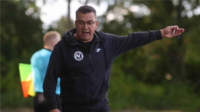 Mann in schwarzer Nike-Jacke mit Kapuze zeigt mit ausgestrecktem Arm nach rechts, unscharfer Linienrichter im Hintergrund mit gelb-roter Fahne.