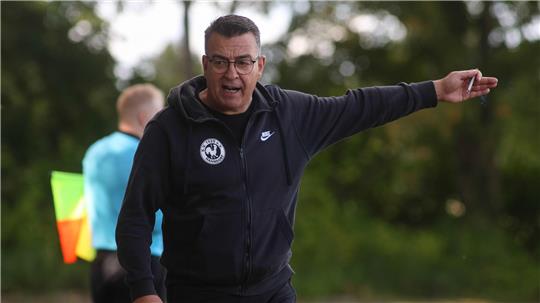 Mann in schwarzer Nike-Jacke mit Kapuze zeigt mit ausgestrecktem Arm nach rechts, unscharfer Linienrichter im Hintergrund mit gelb-roter Fahne.