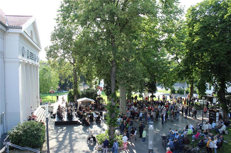 Vor einem repräsentativen weißen Gebäude versammeln sich zahlreiche Menschen entspannt unter Bäumen.