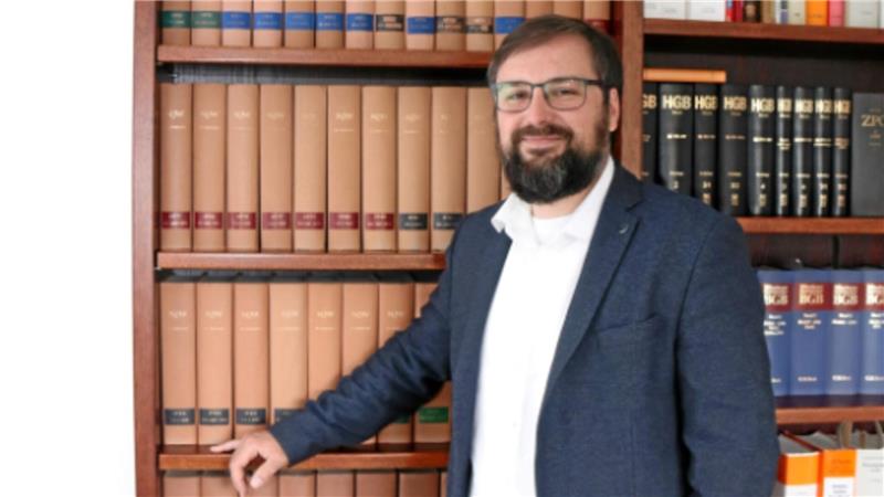 Ein Mann mit Bart trägt ein weißes Hemd und ein dunkelblaues Sakko und steht vor einem Bücherregal mit juristischen Fachbüchern.