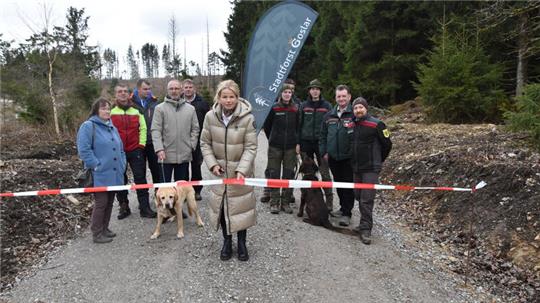 Gruppe von Menschen steht hinter rot-weißem Absperrband auf einem Waldweg, eine Frau in hellem Mantel hält das Band und eine Schere, ein Hund steht vorne links.