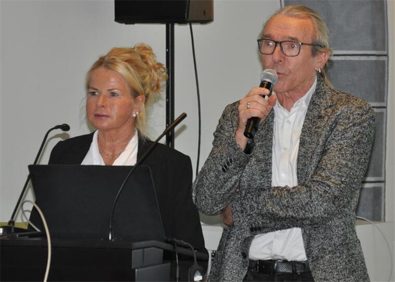 Zwei Personen stehen an einem Pult mit Mikrofonen, eine Person hält ein Mikrofon in der Hand, im Hintergrund ein Lautsprecher und ein Fenster.