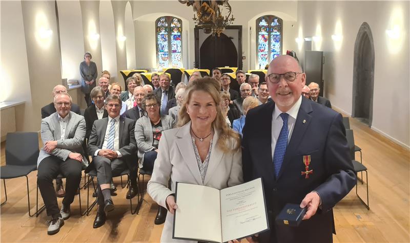 Eine Frau und Ein Mann stehen vor einer Gruppe sitzender Menschen in einem herrschaftlichen Saal mit Holzfußboden. Die Frau hält eine aufgeschlagene Urkunde, der Mann eine Schatulle.