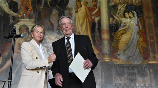 Ein Mann und eine Frau in formeller Kleidung stehen vor einer großen Wandmalerei mit historischen Figuren und Säulen