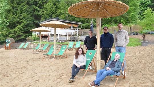 Gruppe von fünf Personen an einem Sandstrand mit Liegestühlen und großen Sonnenschirmen aus Holz und Stroh, umgeben von Bäumen