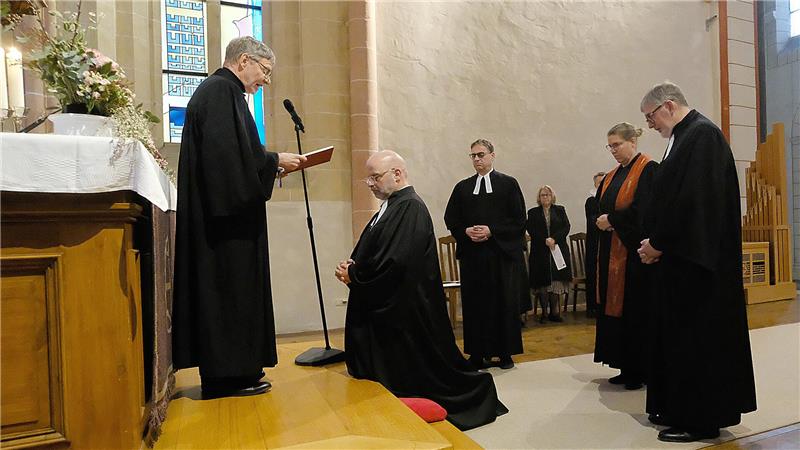 Vor einem Altar kniet ein Mann mit gefalteten Händen. Hinter ihm stehen zwei Männer und eine Frau. Vor ihm steht auf dem Podest ein Geistlicher in schwarzer Robe, der etwas aus einem Buch vorliest. Alle Personen haben schwarze Roben und sind kirchliche Amts- und Würdenträger.