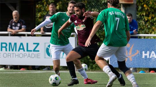 Drei Fußballspieler kämpfen um den Ball auf dem Spielfeld, zwei tragen grüne Trikots, einer ein dunkelrotes Trikot