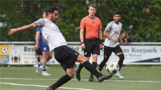 Fußballspieler in weißem Trikot und schwarzer Hose schießt Ball auf Spielfeld mit Schiedsrichter und weiteren Spielern im Hintergrund