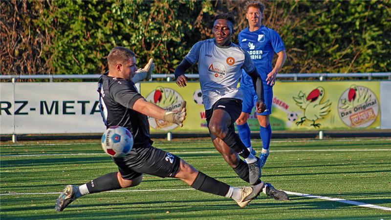 Fußballspieler in Aktion auf dem Spielfeld, ein Spieler in schwarzer Torwartkleidung macht eine Grätsche, während ein Spieler in weißem Trikot den Ball führt und ein Spieler in blauem Trikot im Hintergrund steht