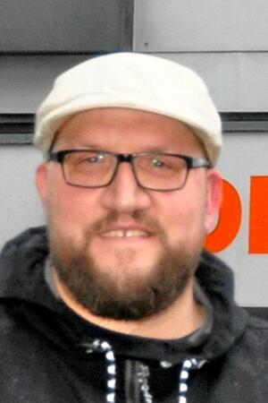 Mann mit heller Kappe, Bart und schwarzer Jacke vor einem Gebäude mit orangefarbenem Schriftzug