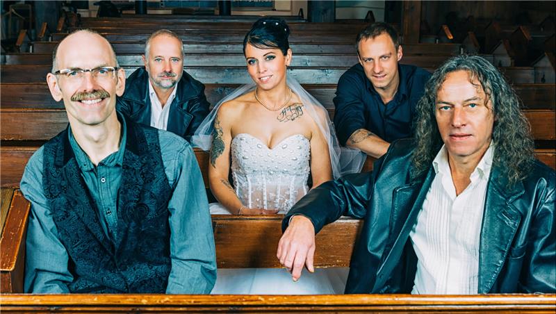 Vier Männer sitzen in einer Kirche, in der Mitte sitzt die Sängerin in einem Brautkleid.