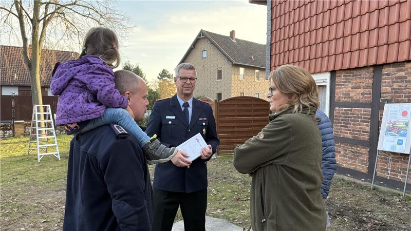 Pferdespritze nimmt ihren Platz im Kleinen Spritzenhaus ein Eine Person in Uniform übergibt ein Dokument an eine Gruppe von Erwachsenen und ein Kind im Freien vor einem Fachwerkhaus.