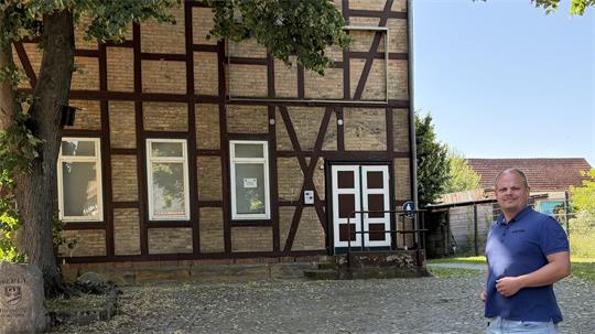Mann in blauem T-Shirt und Jeansshorts steht auf gepflastertem Platz vor einem Fachwerkhaus mit weißen Fenstern und Tür.
