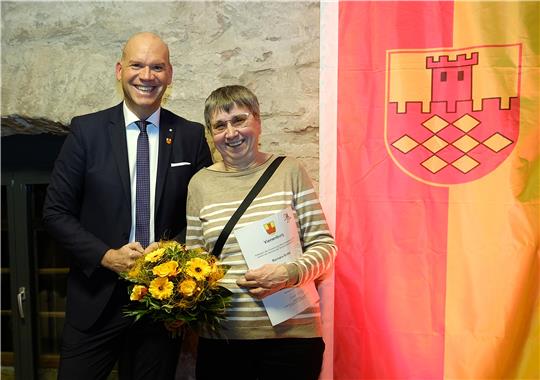 Ein Mann in Anzug und Schlips strahlt in die Kamera, neben ihm lacht eine etwas kleinere Frau mit Brille, Urkunde und gelbem Blumenstrauß etwas gerührt in die Kamera; beide stehen vor einer rot-gelben Fahne mit Burgwappen.