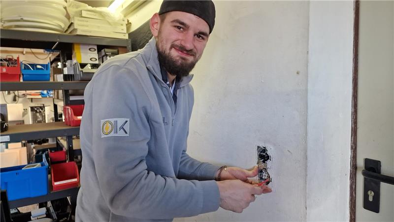 Person in grauem Pullover arbeitet an einer Wandsteckdose in einem Raum mit Regalen und Werkzeugen
