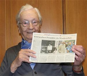Älterer Mann mit Brille hält eine Zeitung mit der Überschrift 'WM beschert Freunden ein Wiedersehen' vor eine Holzwand