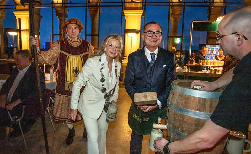 Person in weißem Anzug mit Amtskette hält einen großen Krug neben einem Mann in traditioneller Kleidung mit einem Holzblock vor einem Fass, umgeben von weiteren Personen in einem Innenraum mit großen Fenstern