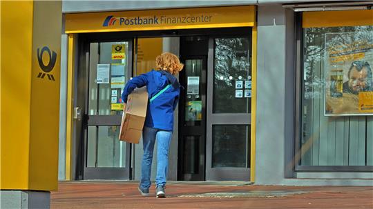 Person mit rotem Haar und blauer Jacke trägt ein Paket auf den Eingang eines Postbank-Finanzcenters zu.