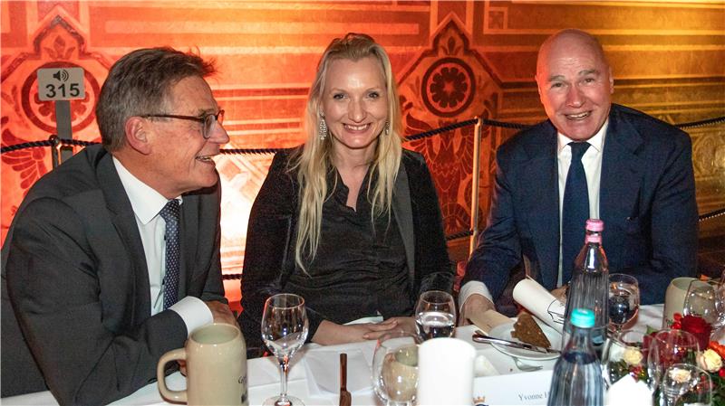 Drei Personen in formeller Kleidung sitzen an einem gedeckten Tisch mit Gläsern, Flaschen und Kerzen in einem warm beleuchteten Raum.