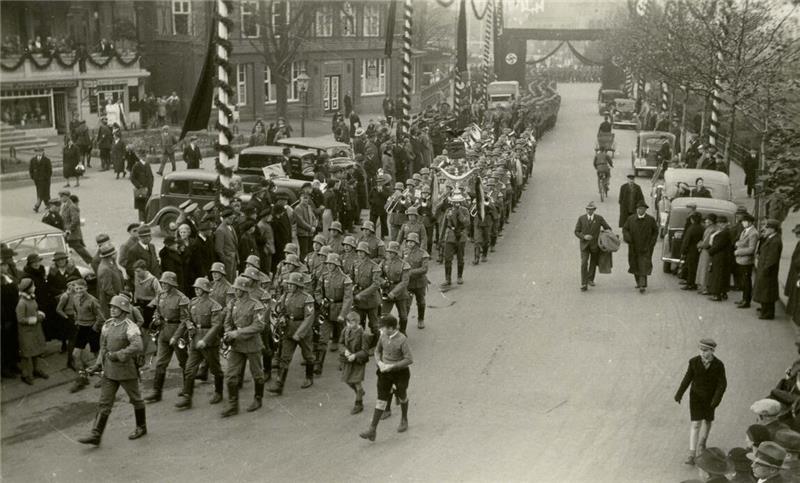 Straßenszene mit einer marschierenden Militärkapelle und Soldaten, umgeben von Zuschauern und parkenden Autos
