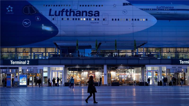Passagiere gehen am Münchner Flughafen vor dem Terminal 2 an einem Plakat mit Flugzeugen der Lufthansa entlang.