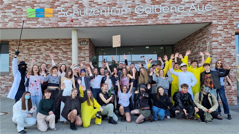 Gruppe von Jugendlichen und jungen Erwachsenen vor dem Gebäude mit Schriftzug 'Schulzentrum Goldene Aue' die in die Kamera winken und lächeln
