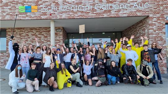 Gruppe von Jugendlichen und jungen Erwachsenen vor dem Gebäude mit Schriftzug 'Schulzentrum Goldene Aue' die in die Kamera winken und lächeln