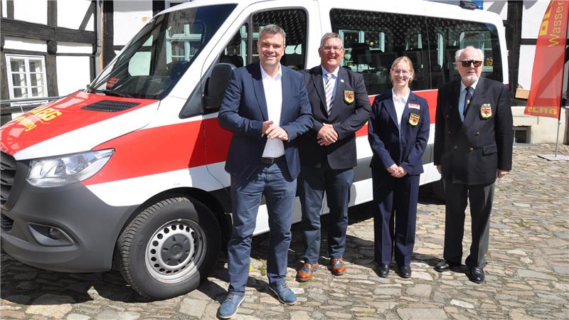 Vier Personen in Anzügen stehen vor einem weißen Fahrzeug mit rotem Streifen auf einem gepflasterten Platz