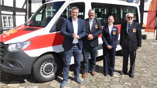 Vier Personen in Anzügen stehen vor einem weißen Fahrzeug mit rotem Streifen auf einem gepflasterten Platz