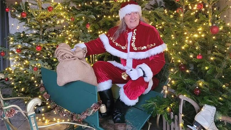 Person in rotem Weihnachtsmannkostüm mit weißem Pelzbesatz sitzt auf grünem Schlitten vor beleuchtetem Weihnachtsbaum, hält großen Sack und trägt schwarze Stiefel.