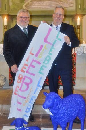 Pastor André Dittmann steht links auf dem Foto, rechts daneben sein Mann Ramon. Beide tragen einen schwarzen Anzug. Gemeinsam halten sie ein Banner, auf dem in bunten Buchstaben die Worte "Liebe" und "Leben" stehen.