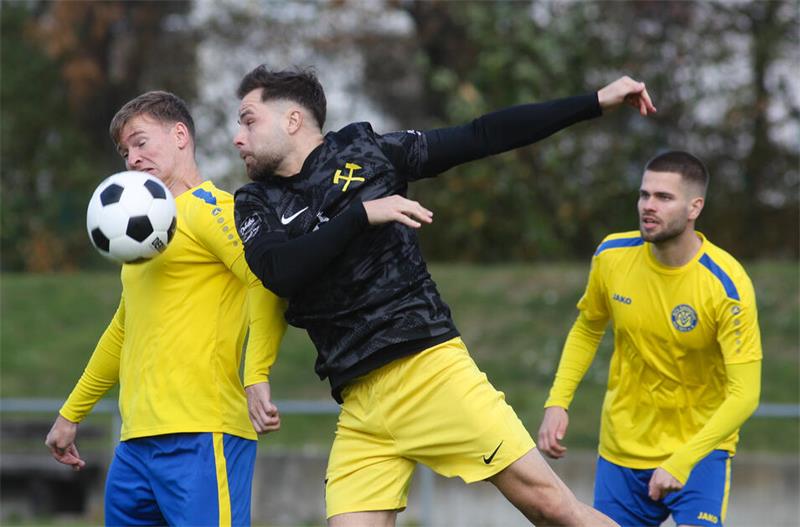 Drei Fußballspieler in gelben Trikots kämpfen um den Ball, einer springt mit ausgestrecktem Arm und Bein hoch