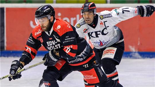 Zwei Eishockeyspieler in Aktion auf dem Eis, einer in schwarzer und roter Ausrüstung, der andere in weißer Ausrüstung.