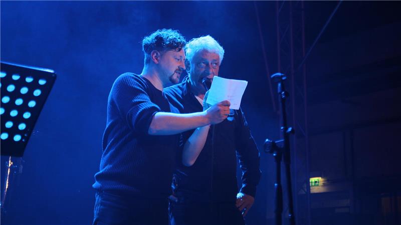 Paul Kunze und Maik Herrmann stehen auf der Bühne und lesen bei der Begrüßung des Publikums abwechselnd die Sponsoren vom einen Zettel vor.