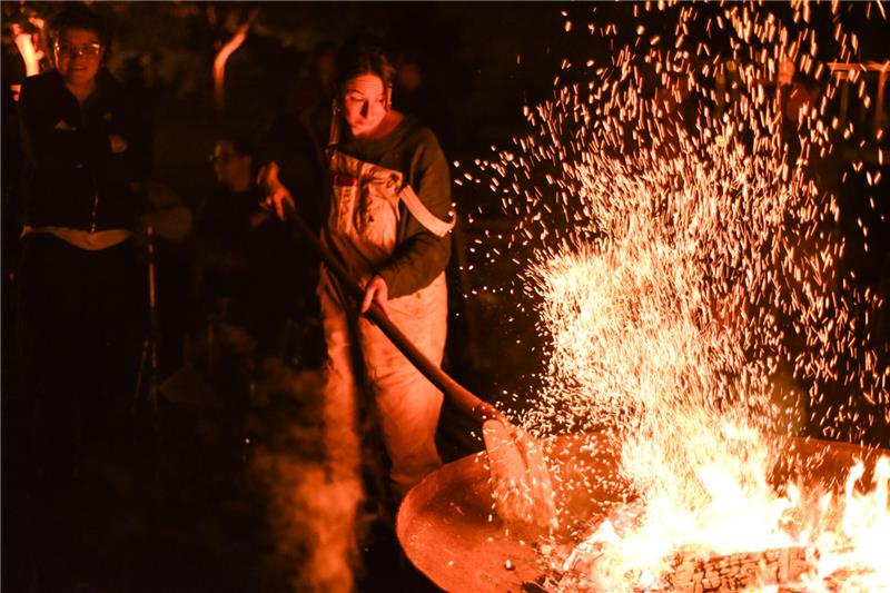 Eine Frau schürt das funkensprühende Feuer in einer großen Feuerschale mit einer Schaufel.