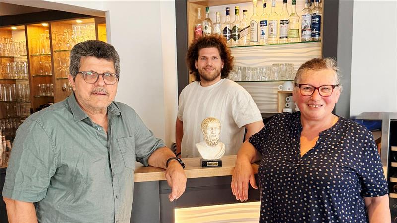 Drei Personen stehen an einer Bar mit Regalen voller Flaschen und Gläser, auf der Theke steht eine Büste eines Mannes