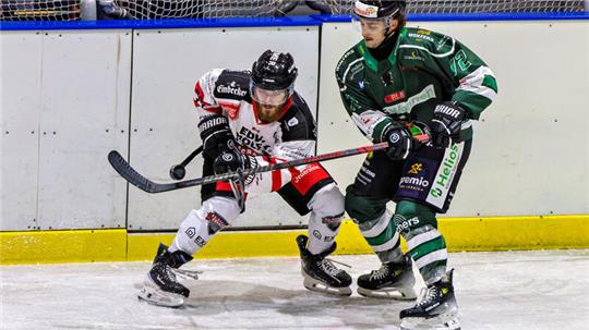 Zwei Eishockeyspieler in Aktion, einer in weiß-roter, der andere in grün-schwarzer Ausrüstung, kämpfen um den Puck an der Bande.
