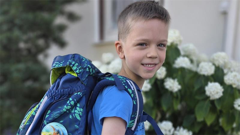 Junge mit blauem T-Shirt und Rucksack steht vor einem Strauch mit weißen Blüten. 