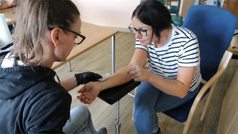Zwei Frauen mit Brillen sitzen an einem Tisch, eine legt ein Blatt Papier auf den rechen Arm der anderen.