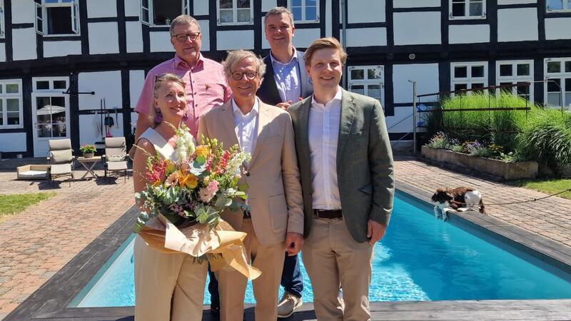 Fünf Personen stehen vor einem Pool und einem Fachwerkhaus, eine Frau hält einen großen Blumenstrauß..
