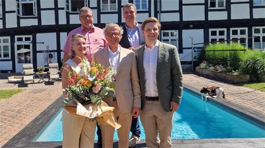 Fünf Personen stehen vor einem Pool und einem Fachwerkhaus, eine Frau hält einen großen Blumenstrauß..