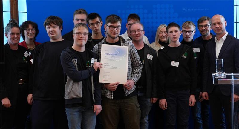 Eine Gruppe von Menschen steht vor blauem Hintergrund, eine Person hält eine Urkunde in der Mitte der Gruppe.