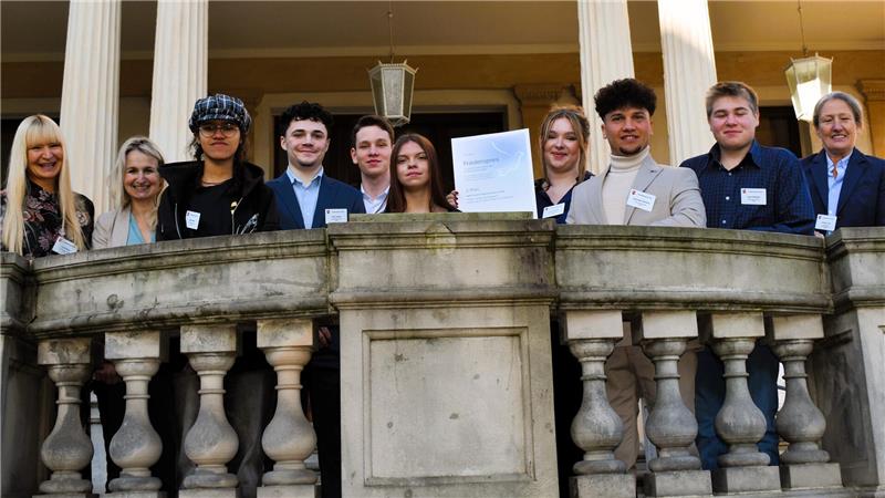 Auszeichnung für Projekte über NS-Opfer und jüdisches Leben Gruppe von Menschen steht hinter einer steinernen Balustrade vor einem Gebäude mit Säulen, eine Person hält eine Urkunde hoch.