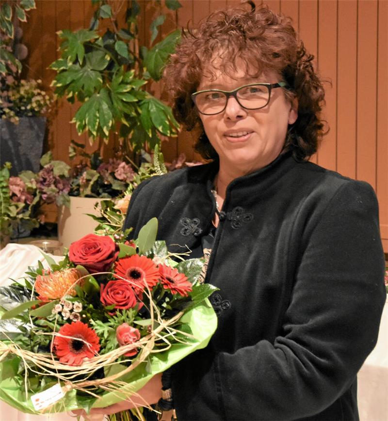 Frau mit lockigem Haar trägt schwarzen Mantel und hält einen Blumenstrauß mit roten Rosen und anderen Blüten vor einem Holzhintergrund und grünen Pflanzen.