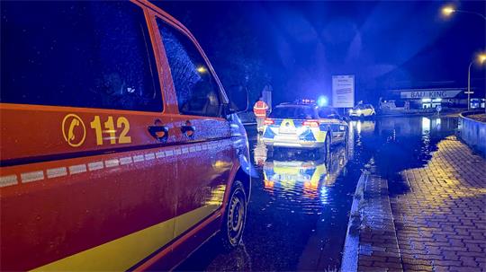 Rotes Einsatzfahrzeug mit der Nummer 112 steht bei Nacht auf nasser Straße neben einem Wasserpfütze, im Hintergrund weitere Einsatzfahrzeuge mit Blaulicht