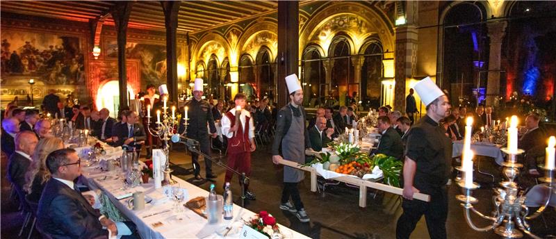Festlich gekleidete Gäste sitzen an langen Tafeln in einem prunkvollen Saal mit hohen Bögen und Kerzenleuchtern, während Köche mit weißen Hüten ein großes Tablett mit Speisen tragen.