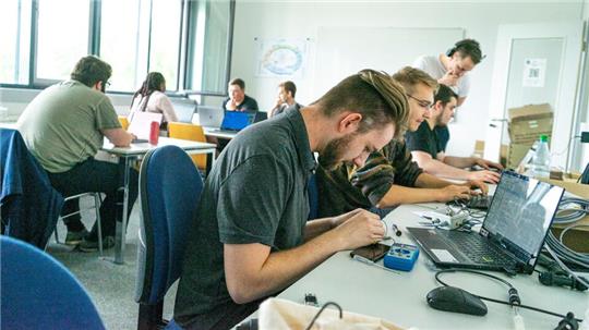 Das Foto zeigt sieben Studenten am Tisch mit Laptops und Elektronik bei der Projektarbeit. Ein Dozent ist stehend mit ihnen im Gespräch.